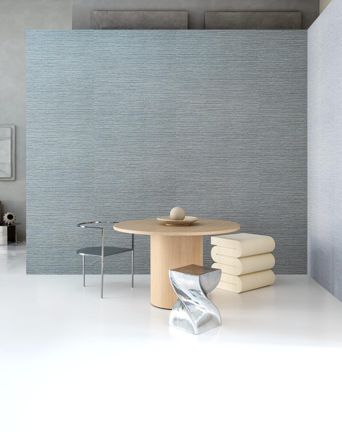 A l'arrière plan, un revêtement mural tissé bleu-gris, avec devant une table en bois clair, une chaise en métal et deux tabourets, un en métal et l'autre de couleur beige. Le sol est blanc.